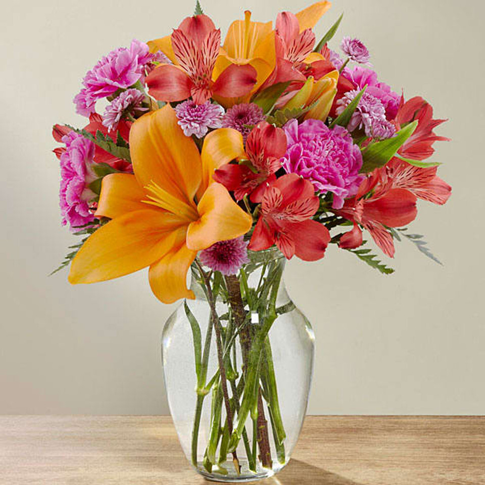 A cheerful bouquet of bright orange lilies, deep pink carnations, red Peruvian lilies, and soft lavender chrysanthemums, beautifully arranged in a clear glass bubble bowl vase.