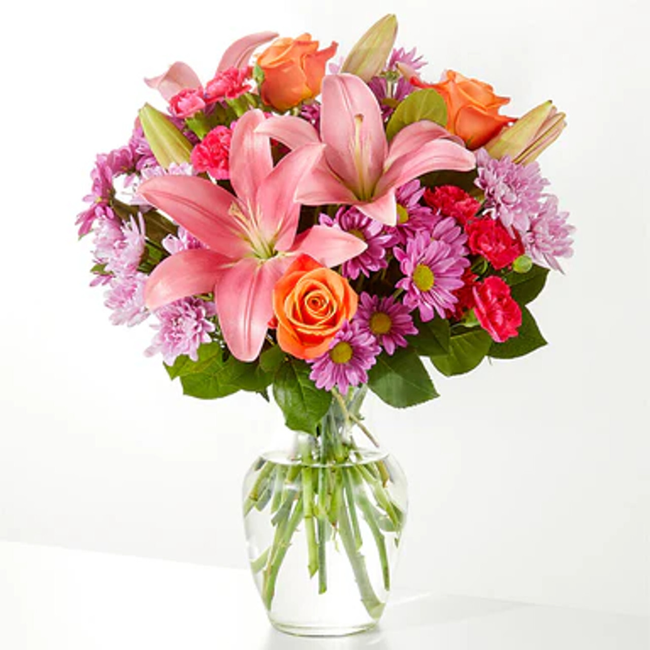 A vibrant floral arrangement in a clear glass vase, featuring pink lilies, orange roses, purple daisies, red carnations, and lush green foliage.