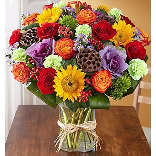 Vibrant bouquet with sunflowers, red roses, purple lisianthus, green carnations, and berries in a twine-tied glass vase.