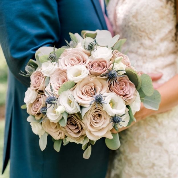 Close-up wedding bouquet with dusty-rose and cream roses, white lisianthus, eucalyptus, and blue thistles.