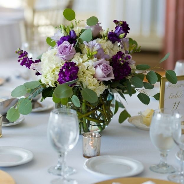 Wedding table decor featuring a glass vase of purple and white flowers with greenery, surrounded by plates and water glasses.