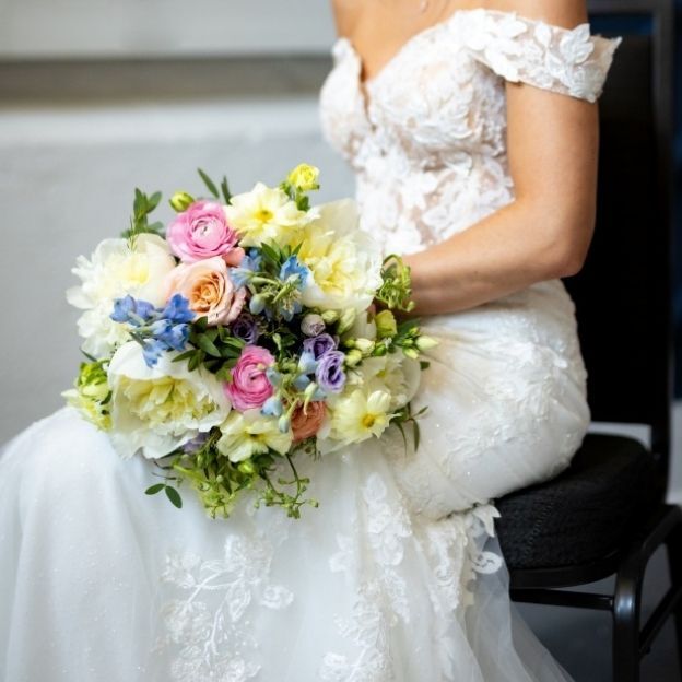 Close-up of a seated bride in a lace dress with a vibrant bouquet featuring peonies, roses, and mixed spring flowers.
