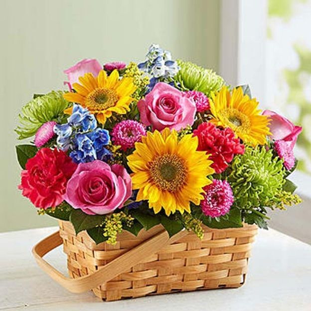 Bright flower arrangement in a wooden basket featuring sunflowers, roses, carnations, mums, and blue delphiniums.