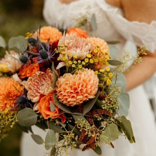 Close-up of a wedding bouquet with orange and peach flowers, yellow accents, and lush green leaves.
