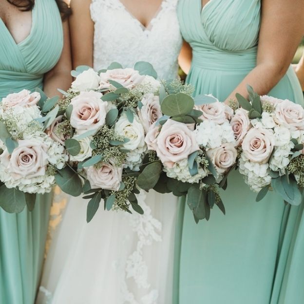 Three women in mint green dresses and a white lace gown hold bouquets of pale pink roses, white flowers, and greenery.