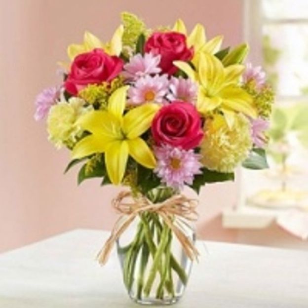 Bright floral bouquet with yellow lilies, pink daisies, and red roses arranged in a glass vase with a twine bow.