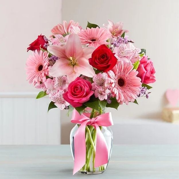 Clear glass vase with pink gerberas, roses, alstroemeria, and a pink lily, finished with a pink ribbon bow.