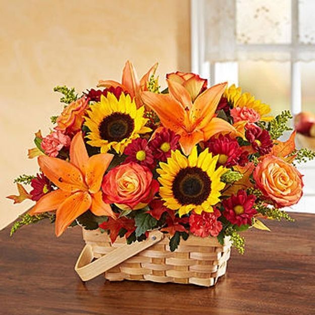 Fall basket blends sunflowers, orange Asiatic lilies, mini carnations, bi-color or circus roses, and red daisy poms in a split-wood handled basket