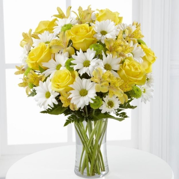 Tall glass vase with yellow roses, white daisies, yellow alstroemeria, green button mums, and ferns by a window.
